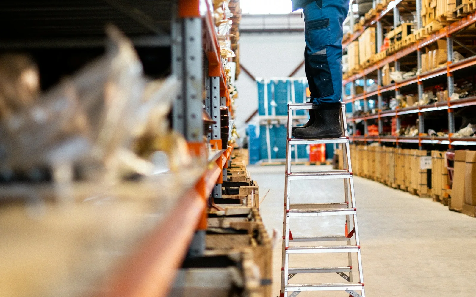 Warehouse worker coordinating inventory from a ladder between stocked shelves